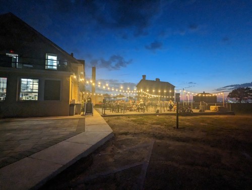 A cozy outdoor dining area illuminated by string lights at dusk, with a building in the foreground and a clear blue sky.
