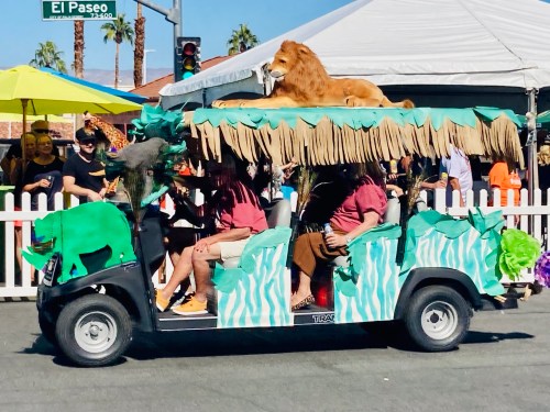A decorated golf cart with a lion on top, surrounded by people in tropical attire, driving through a festive parade.