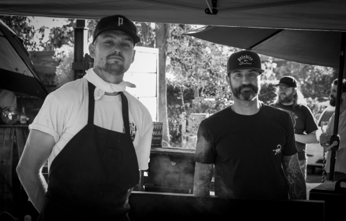 Two chefs in black and white, one wearing a cap and apron, the other with a beard, standing behind a food stall.