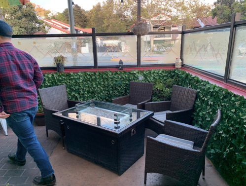A cozy outdoor seating area with a glass-top table and wicker chairs, surrounded by greenery.