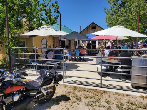 Outdoor dining area with umbrellas, people seated at tables, and a motorcycle parked nearby. Bright, sunny day.