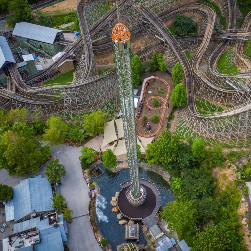 Aerial view of a tall amusement park drop tower surrounded by roller coasters and greenery.