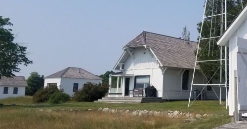 A white house with a porch and a black grill, surrounded by grass and trees, with another building in the background.