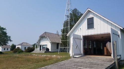 Two white buildings with a tall tower, surrounded by grass and trees, on a clear day. One building has an open garage.