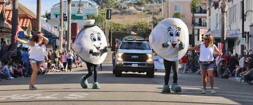 Two large clam mascots parade down the street, accompanied by two women, with a crowd watching in the background.