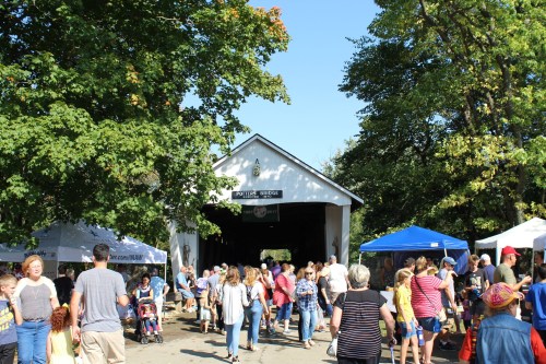 A busy covered bridge with people walking, surrounded by trees and tents at a lively outdoor event.