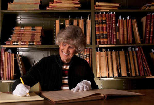 An elderly woman in a black sweater smiles while writing in a large book, surrounded by shelves of old books.