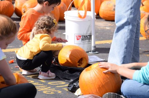 Children carving pumpkins at an outdoor event, with a bucket and scattered pumpkins in the background.