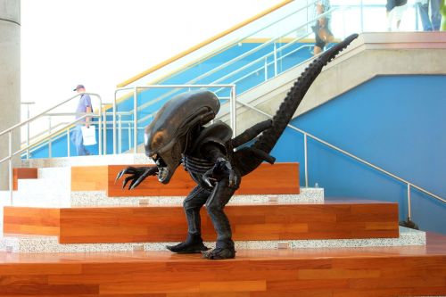 A person in a detailed alien costume poses on a staircase in a modern building.