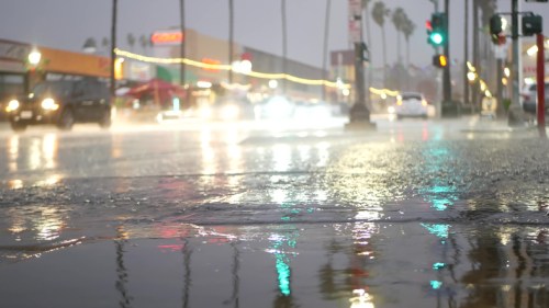 Rain-soaked street with reflections, cars driving by, and traffic lights glowing in a stormy atmosphere.