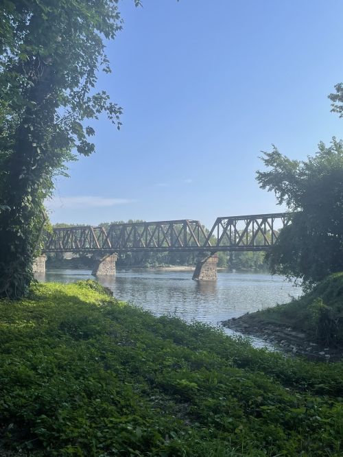 A rusted railway bridge spans a calm river, surrounded by lush greenery and a clear blue sky.