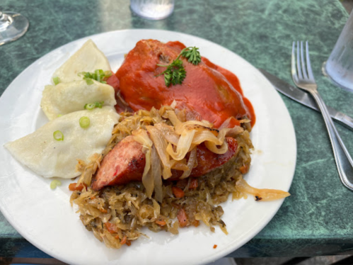 A plate of sausage with sauerkraut, topped with sauce, alongside dumplings and garnished with parsley.