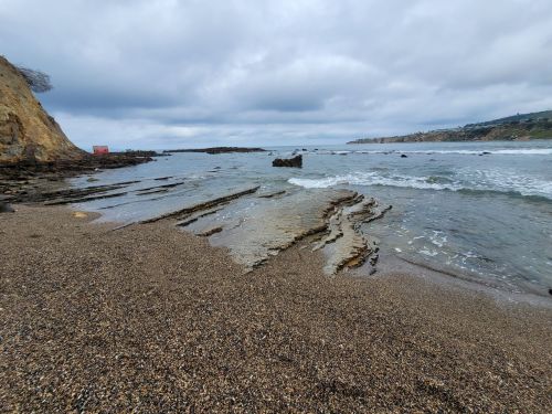 A rocky beach with gentle waves under a cloudy sky, featuring layered rock formations and a distant coastline.