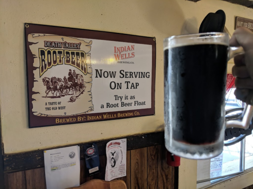 A hand holds a frosty mug of root beer in front of a sign announcing it’s now on tap at Indian Wells Brewing Co.