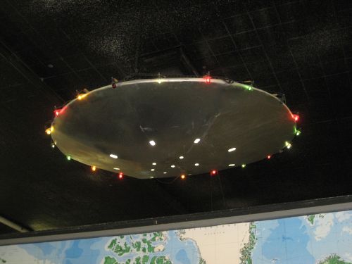 A metallic, circular object resembling a UFO, adorned with colorful lights, suspended from a ceiling above a map.