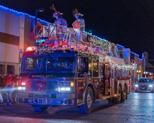 A decorated fire truck with lights and two figures in reindeer antlers, parading at night.