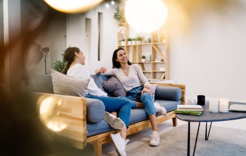 Two women sit on a couch, laughing and chatting in a cozy, modern living space with plants and soft lighting.