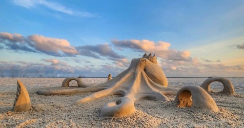 A large sand sculpture of an octopus on a beach, with a colorful sky and clouds in the background.