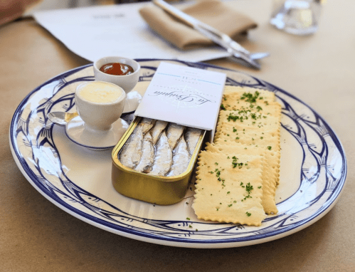 A plate with a tin of sardines, crackers, a small cup of butter, and a side of sauce, garnished with parsley.
