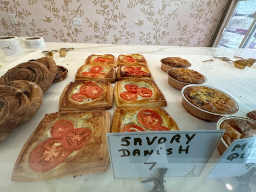 A display of pastries, including savory danishes topped with tomatoes and sweet muffins, in a bakery setting.