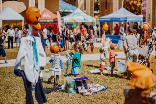 A lively outdoor event with children playing, pumpkin decorations, and colorful tents in the background.