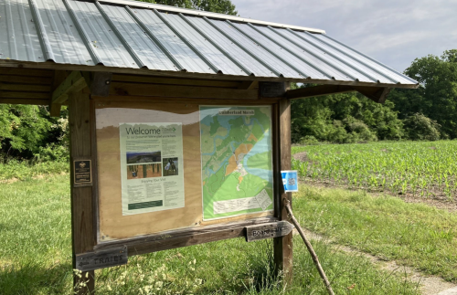 A wooden information board with a map and welcome sign, set in a grassy area near a field.