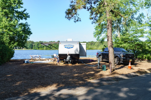 A camper parked by a lake, surrounded by trees, with a truck nearby and a picnic area in the background.