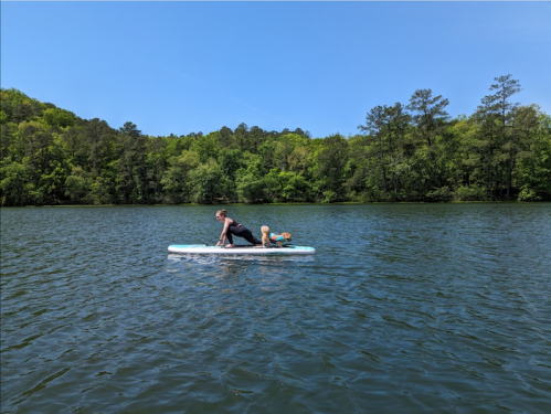 A person paddles on a stand-up paddleboard with a child sitting behind them on a calm lake surrounded by greenery.