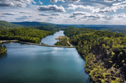 Aerial view of a serene lake surrounded by lush green hills and a winding road under a partly cloudy sky.