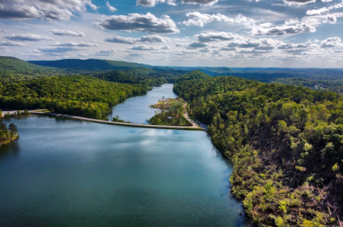 Aerial view of a serene lake surrounded by lush green hills and a winding road under a partly cloudy sky.