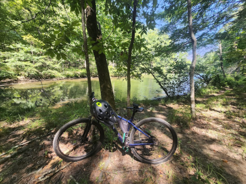 A mountain bike with a helmet rests by a serene river, surrounded by lush green trees and sunlight.