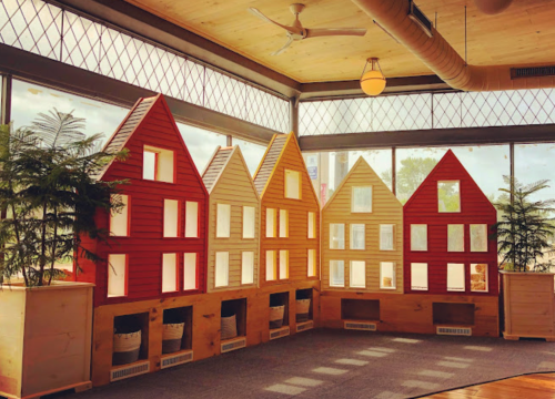 Colorful wooden house-shaped structures with windows, arranged in a bright, airy indoor space with plants.