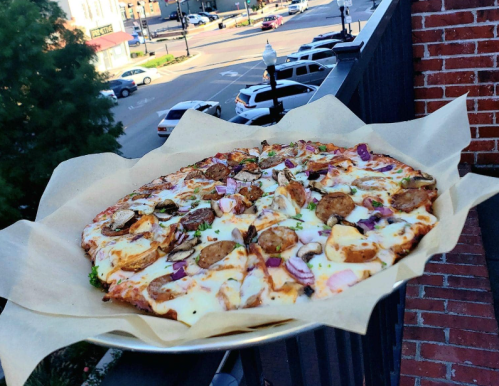 A freshly baked pizza topped with mushrooms, onions, and herbs, served on a paper-lined tray with a city view in the background.