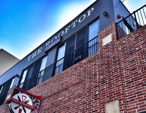 A brick building with a sign reading "The Rooftop" and "Open," featuring a balcony and clear blue sky above.