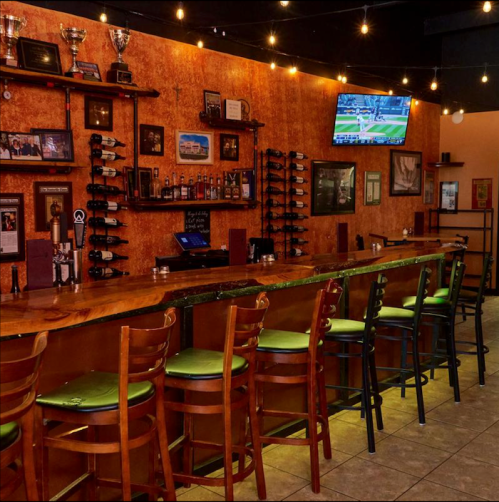 A cozy bar with wooden counters, green stools, trophies on the wall, and a TV displaying a sports game.