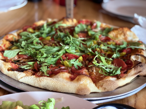 A freshly baked pizza topped with tomatoes, peppers, and arugula on a metal tray, with a salad in the foreground.