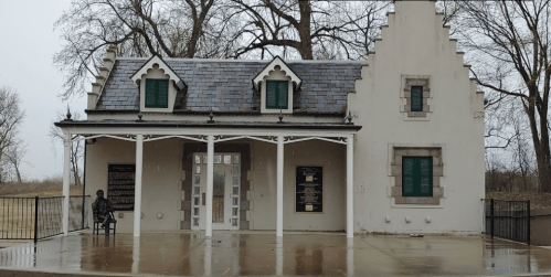 Historic building with green shutters, a porch, and a statue seated outside, surrounded by bare trees on a rainy day.