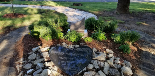 A heart-shaped memorial stone surrounded by greenery and rocks, set in a landscaped area.