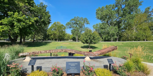 A metal sculpture on a stone base in a park, surrounded by greenery and flowers, with plaques nearby.