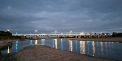A river at dusk with a bridge illuminated by lights reflecting on the water's surface.