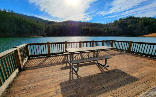 A wooden dock with a picnic table overlooks a serene lake surrounded by trees and mountains under a blue sky.