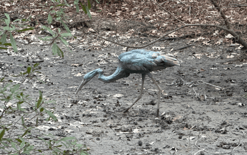 A blue heron statue wades through a muddy area surrounded by dry leaves and sparse vegetation.