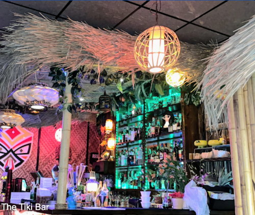 A vibrant tiki bar with tropical decor, colorful lights, and a well-stocked bar featuring various drinks and plants.