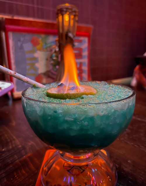 A vibrant blue cocktail with a flaming garnish and a slice of lime on top, served in a decorative glass.