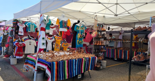 A vibrant market stall featuring colorful clothing, handmade shoes, and traditional textiles under a white canopy.