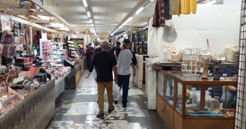 A bustling market aisle with shoppers, surrounded by various stalls selling goods and merchandise.