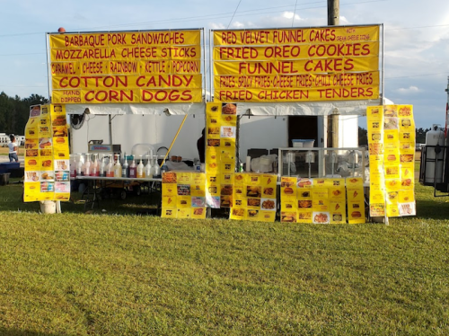Food vendor stand with colorful signs advertising BBQ pork, corn dogs, funnel cakes, and various snacks.