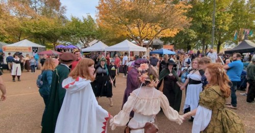A lively outdoor festival scene with people in costumes dancing in a circle, surrounded by tents and autumn foliage.