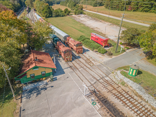 Aerial view of a train station with vintage locomotives, a red caboose, and green fields in the background.