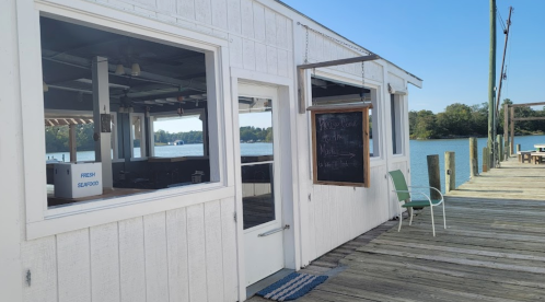 A waterfront seafood restaurant with white siding, a chalkboard sign, and a wooden dock extending into the water.